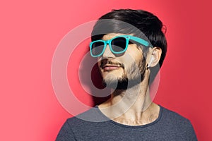 Studio portrait of young cheerful man, wearing cyan sunglasses and using wireless earphones on coral pink background.