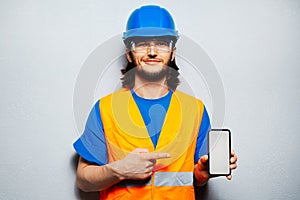 Studio portrait of young construction worker engineer wearing safety equipment, pointing finger at smartphone with mockup.