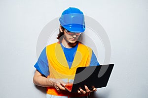 Studio portrait of young construction worker engineer wearing safety equipment, using laptop on background of grey wall.