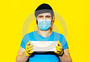 Studio portrait of young delivery man in blue shirt, wearing medical face mask and yellow gloves against coronavirus.