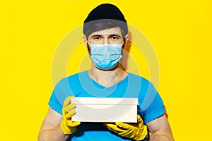 Studio portrait of young delivery man in blue shirt, wearing medical face mask and yellow gloves against coronavirus.
