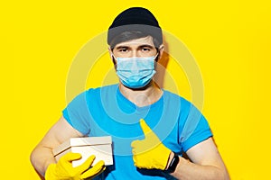Studio portrait of young delivery man in blue shirt, wearing medical face mask and yellow gloves against coronavirus.