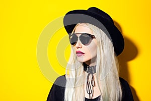 Studio portrait of young fashion blonde hipster girl with pink lips, wearing sunglasses, black hat and choker, isolated on yellow