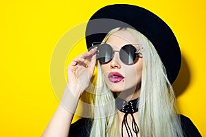 Studio portrait of young fashion blonde hipster girl with pink lips, wearing sunglasses, black hat and choker, isolated on yellow.