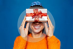 Studio portrait of young girl holding white gift box with red bow on eyes. Background of blue color. Wearing orange sweater.