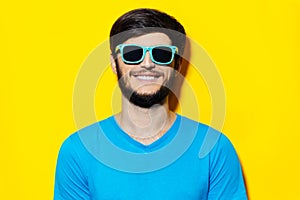 Studio portrait of young guy in blue shirt, wearing cyan sunglasses on background of yellow color.