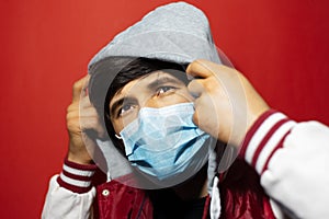 Studio portrait of young guy wearing medical flu mask, coronavirus protect, puts on hood of jacket, looking up on red background.