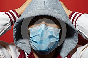 Studio portrait of young hooded guy wearing medical flu mask, coronavirus protect, holding hands on head on red background.