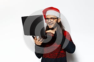 Studio portrait of young laughing man in red sweater, holding laptop, typing on keyboard of computer, wearing Santa Claus hat.