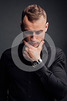 Studio portrait of young man in black shirt, holding hands in chin, looking down with humility.