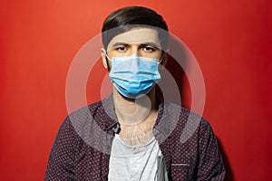 Studio portrait of young man wearing medical face mask against coronavirus on red background.