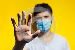 Studio portrait of young man wearing medical flu mask on background of yellow color. Close-up of male hand showing stop gesture.