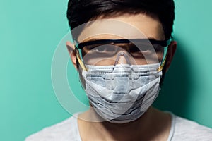 Studio portrait of young man wearing medical flu mask and safety goggles against coronavirus. On aqua menthe background.