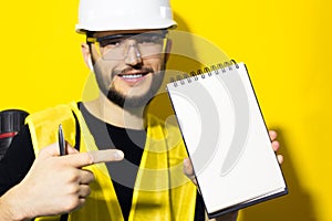 Studio portrait of young modern smiling man architect, showing with finger on notebook with white mockup. Wearing construction saf