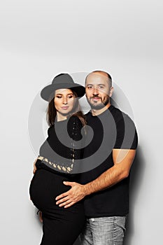 Studio portrait of young parents, pregnant woman wearing black hat and dress, man touching the belly on white background.