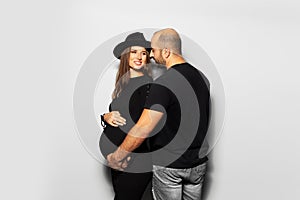 Studio portrait of young parents, pregnant woman wearing black hat and dress, man touching the belly on white background.