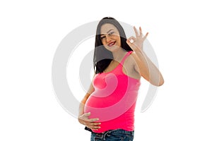 Studio portrait of young pregnant brunette woman in pink shirt touching her belly and showing OK on camera isolated on
