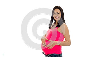 Studio portrait of young pregnant brunette woman in pink shirt touching her belly and smiling on camera isolated on