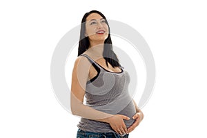Studio portrait of young pregnant brunette woman in shirt touching her belly and looking up and smiling isolated on