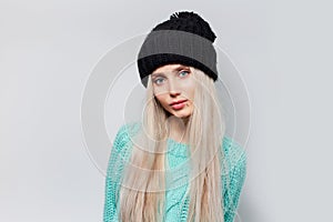Studio portrait of young pretty blonde girl on background of white textured wall, wearing blue sweater and black hat.