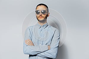 Studio portrait of young serious man with crossed arms, wearing sunglasses and blue shirt on white background