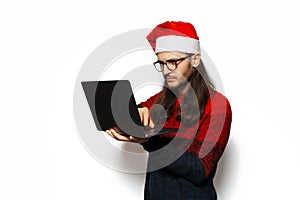 Studio portrait of young serious man in red sweater, holding laptop, typing on keyboard of computer, wearing Santa Claus hat.