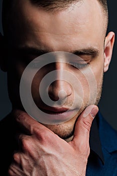 Studio portrait of young man in black shirt, holding hands in chin, looking down with humility.