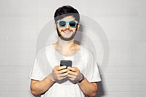 Studio portrait of young smiling guy with smartphone in hands, wearing cyan shades, background of white color.