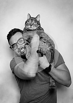 Studio portrait of a young smiling man with a beard and glasses holding a tabby cat in his arms. Grey background. White background