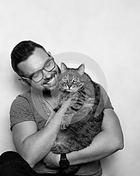 Studio portrait of a young smiling man with a beard and glasses holding a tabby cat in his arms. Grey background. White background