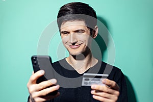 Studio portrait of young smiling man shopping online by smartphone and credit card on background of aqua menthe color.