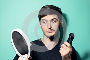 Studio portrait of young surprised man with half shaved face, looking in mirror holding electric shaver trimmer on background.