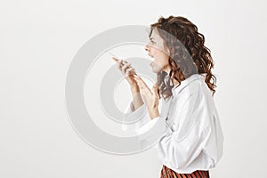 Studio portrait of young trendy woman standing in profile shouting at smartphone while holding gadget in hand, gesturing