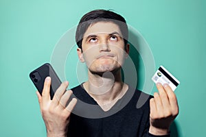 Studio portrait of young upset man looking up holding in hands smartphone and credit card on background of aqua menthe color.
