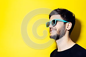 Studio profile portrait of young smiling man looking away, wearing cyan sunglasses on yellow background with copy space.