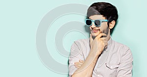 Studio profile portrait of young thoughtful guy wearing cyan sunglasses, holding hand on chin, looking on empty background of cyan