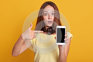 Studio sho of adorable woman showing phone with blank screen and pointing on it with her index finger, has astonished facial