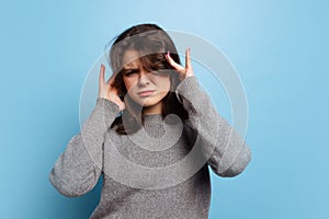 Studio shot of angry young beautiful girl in warm gray knitted sweater isolated on blue color background