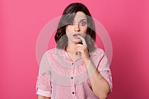 Studio shot of attractive european female with dark hair, holding her finger on lower lip over rose background, looking directly