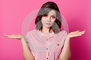 Studio shot of attractive woman shrugging helpless with her hands, wearing stylish stripped shirt, looking at camera with sad