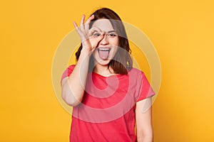 Studio shot of attractive young woman, makes ok sign and covers her eye, shows tongue, expresses happyness, model poses against