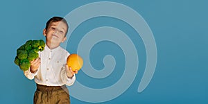 A studio shot of a boy holding fresh broccoli and an orange on a blue background with a copy of the space. The concept