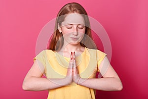 Studio shot of calm beautiful Caucasian teenager girl stands isolated over pink background while praying, keeps eyes closed, asks