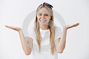 Studio shot of careless calm and carefree young woman with blond hair shrugging with raised palms aside and smiling