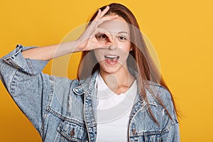 Studio shot of charming young woman, female making ok sign and covers her eye, posing with open mouth, expresses happyness, model