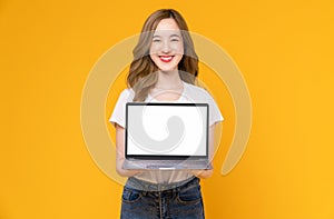 Studio shot of cheerful beautiful Asian woman in white t-shirt and holding laptop mockup of blank screen on yellow background