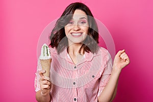 Studio shot of cheerful dark haired woman holds ice cream, clench fist, has happy facial expression, posing in striped shirt, has