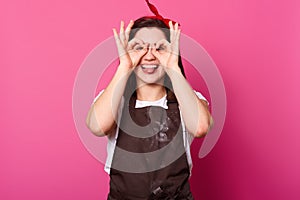 Studio shot of cheerful smiling young woman, covering her eyes with ok signs, expresses joy, model poses against pink background,