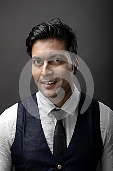 Studio shot of cheerful, young, handsome Indian male model in formal wear wearing white shirt, black tie, black coat against grey