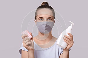 Studio shot of confident serious cute young female holding disinfectant alcohol gel and soap, sticking to rules, protecting